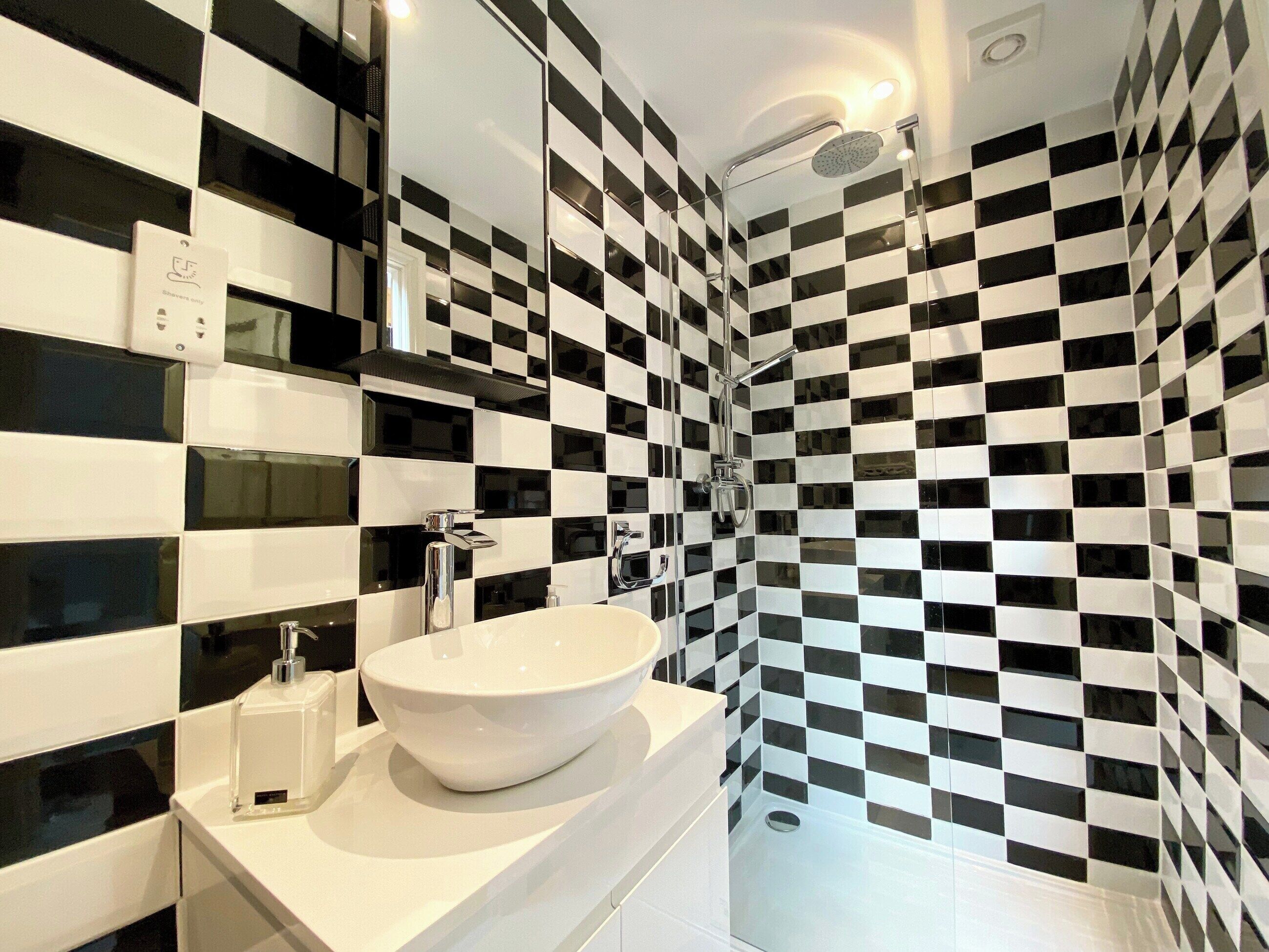 A modern bathroom with a checkered black and white tile wall, a white sink with a soap dispenser, and a shower area with a handheld showerhead.