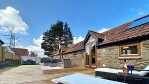 Un terrazzo in legno con mobili da esterno, un edificio in pietra con tetto in tegole e un albero di grandi dimensioni.