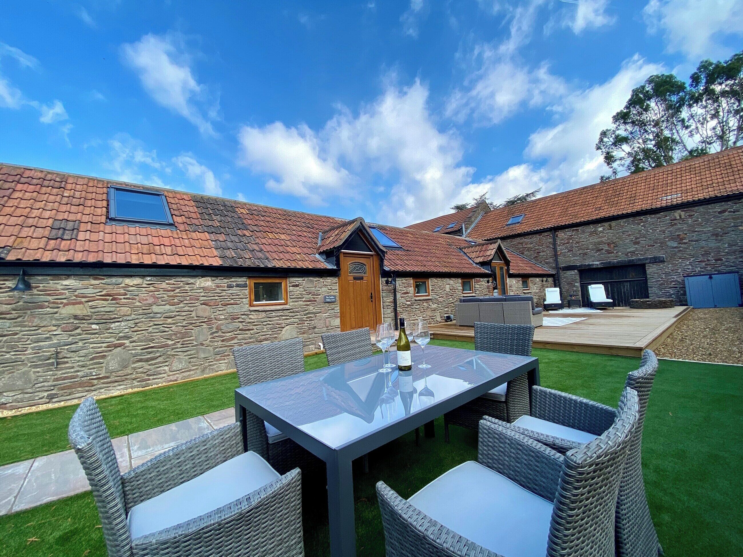 A stone building with a tiled roof, a glass table with chairs, and a grassy lawn.