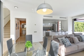Un soggiorno moderno con un tavolo da pranzo in vetro, un divano con cuscini decorati e una cucina sullo sfondo.