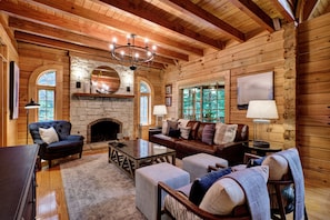 A log cabin interior with a stone fireplace, wooden beams, a chandelier, and a cozy seating area. - Needlepine Lodge of Sapphire Valley, Needlepine Lodge of Sapphire Valley (Jackson County)