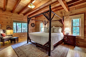 A wooden cabin bedroom with a four-poster bed, a wooden bench, a small table with a lamp, and a window with a view of the outdoors. - Needlepine Lodge of Sapphire Valley, Needlepine Lodge of Sapphire Valley (Jackson County)