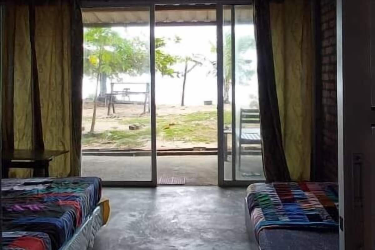A room with two beds, a concrete floor, and a view of an outdoor area with trees and a table.