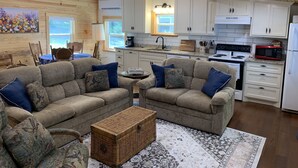 A cozy living room with a sectional sofa, a coffee table, and a wicker chest. Adjacent is a kitchen with white cabinets, a stove, and a microwave. - Lake Cabin with Fantastic View--Kayak, Swim or Fish (Perryville)
