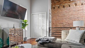 A living room with a sofa, a TV, a plant, and a brick wall. - DC’s Southwest and Navy Yard Welcomes You! (Washington)