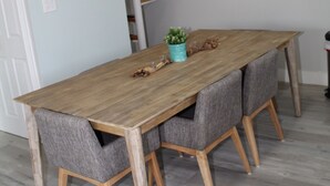 Een eetruimte met een houten tafel, grijs gestoffeerde stoelen, een kamerplant, een kroonluchter, een ronde spiegel en een trap met een witte leuning.