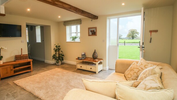 A spacious living room with a large sofa, a wooden coffee table, and a potted plant. There is a television mounted on the wall and a window with a view of the outdoors. - Cow'ouse (Shrewsbury)