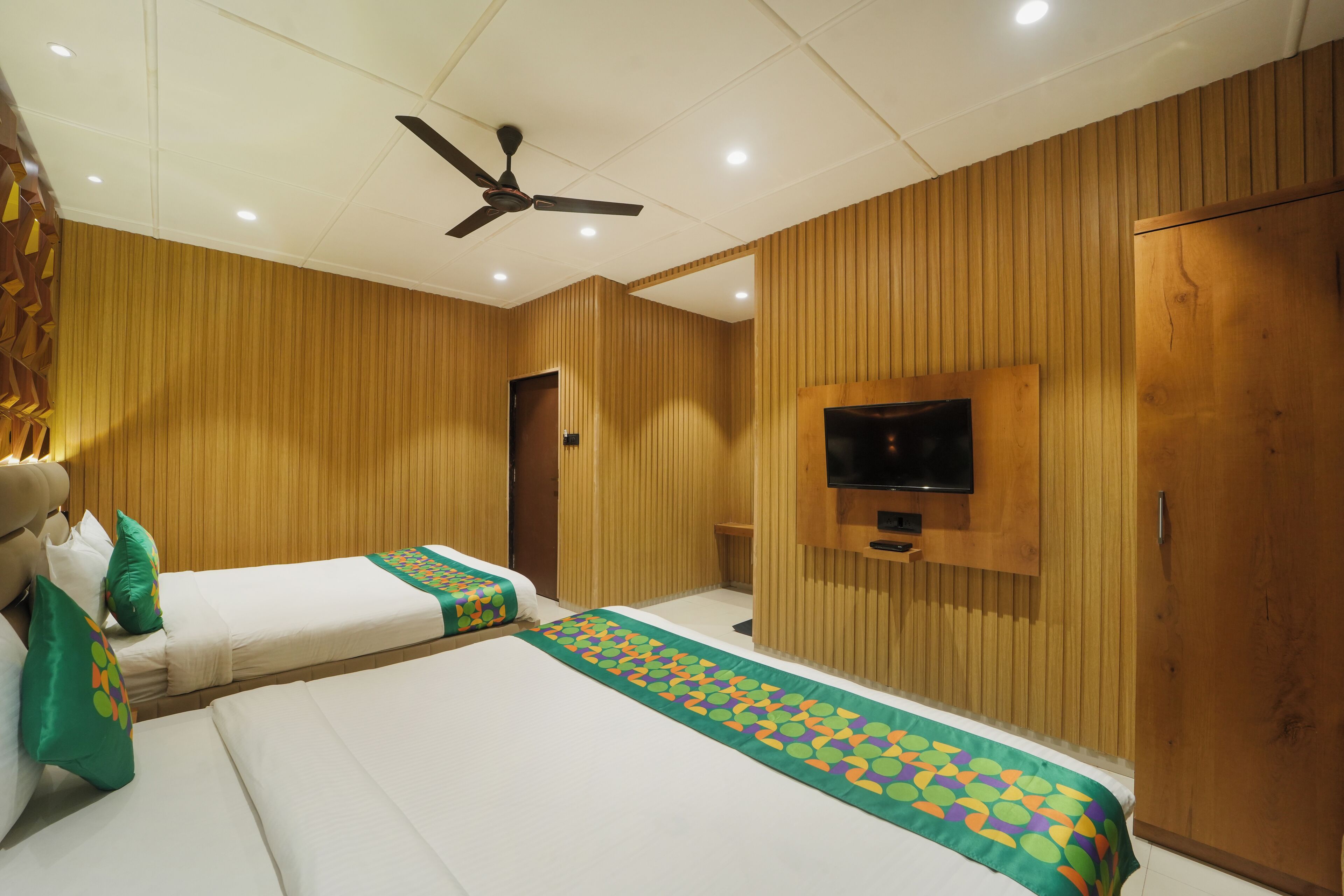A hotel room with two beds, a ceiling fan, a television mounted on the wall, and wooden paneling on the walls.