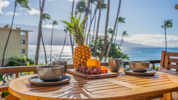 A wooden table with a tropical fruit arrangement and two metal cups, overlooking a beach with palm trees and ocean views.