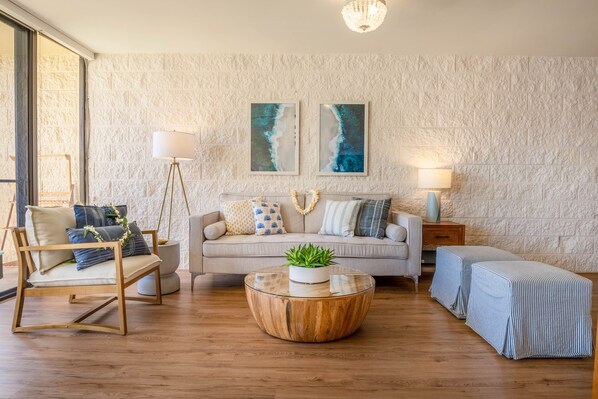 Een woonkamer met een bank, twee fauteuils, een salontafel en een bijzettafeltje. Er zijn twee wandlampen en twee ingelijste foto's aan de muur.