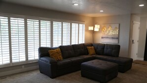 A living room with a sectional sofa, ottoman, large windows with blinds, a floor lamp, and a painting on the wall. - Updated 4-bedroom home centrally located in Logan (Logan)