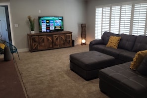 Una sala de estar con sofá modular, reposapiés, mueble de entretenimiento y un televisor de pantalla plana grande que muestra servicios de transmisión.