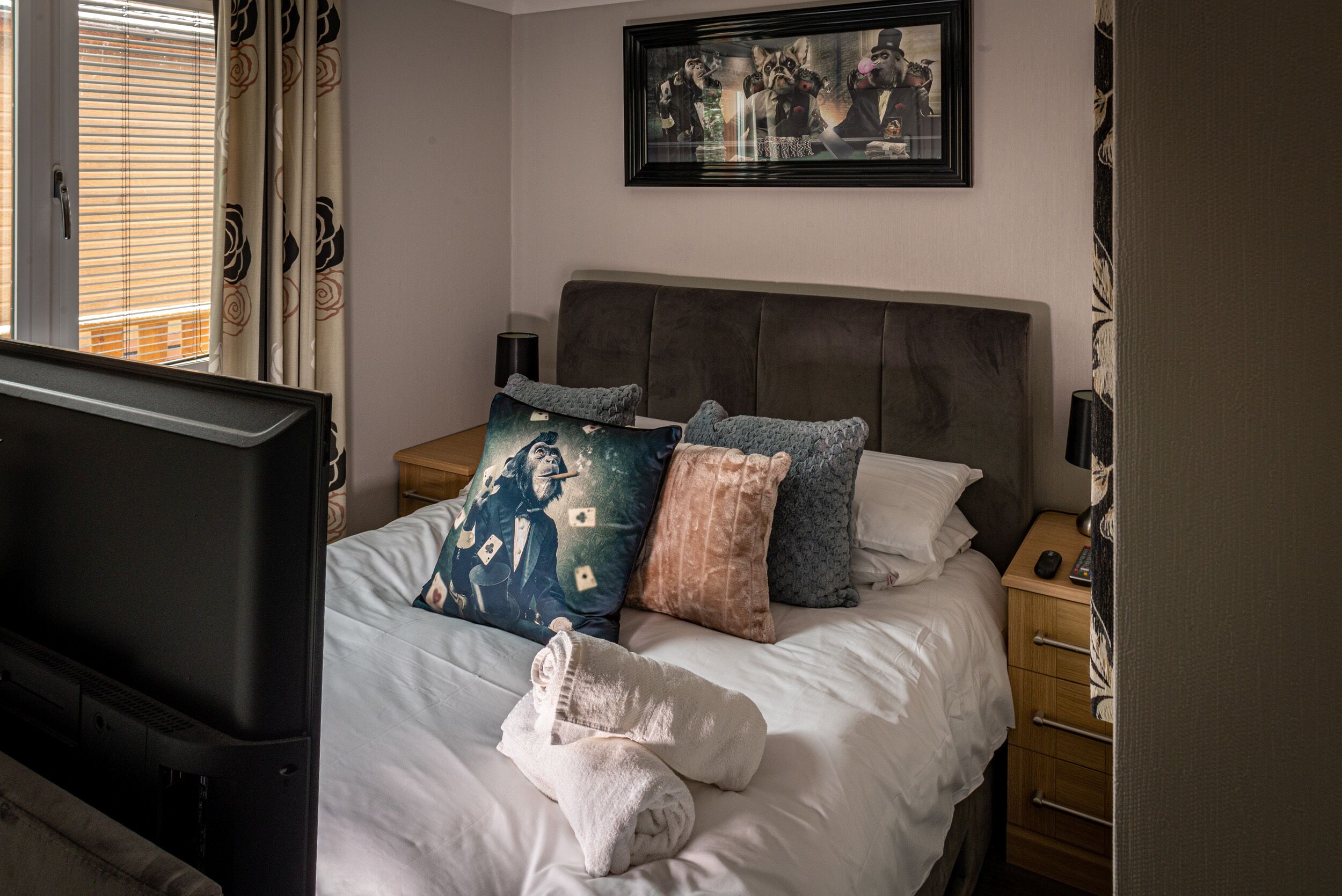 Una cama bien hecha con varias almohadas, un televisor a la izquierda y un cuadro en la pared.