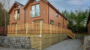 A two-story wooden cabin with a balcony, stone foundation, and a gravel driveway. - Lakeland Lodge (Carnforth)