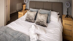 A neatly made bed with a grey knitted blanket, two fluffy pillows, and a rolled towel on the bed. There are two bedside tables with ring lights and a wooden nightstand with a lamp. - Lakeland Lodge (Carnforth)