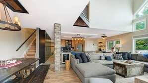 Ein geräumiger Wohnbereich mit einem großen, grauen Ecksofa, einem Holzesstisch und einer Steinsäule. Im Hintergrund ist eine Küche mit modernen Geräten zu sehen.