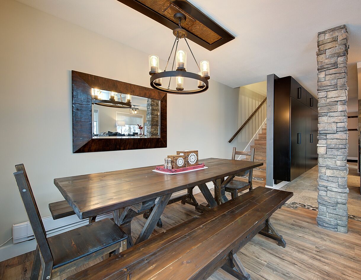 Un espace repas avec une table et des bancs en bois, un lustre, un pilier en pierre et, au fond, une cuisine moderne.