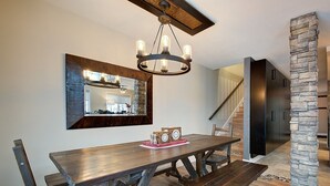 Ein Essbereich mit Holztisch und Bänken, einer Kristallleuchter, einer Steinsäule und einer modernen Küche im Hintergrund.