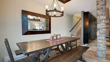 Un espace repas avec une table et des bancs en bois, un lustre, un pilier en pierre et, au fond, une cuisine moderne.