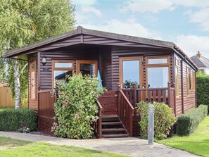 A wooden cabin with a porch, a staircase, and a small garden area. - Lodge Two, Country View Park (Faversham)