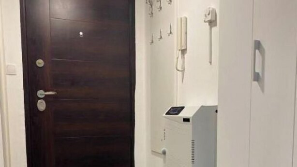 Un couloir avec une porte en bois sombre, un radiateur blanc fixé au mur et un meuble blanc doté d’un panneau de commande.