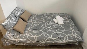 Un petit lit, bien fait, avec un motif abstrait en nuances de gris et de blanc, deux oreillers et une serviette enroulée, le tout placé devant un mur uni.