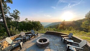 Un patio de piedra con sillas Adirondack y un hogar para fuego, rodeado por un muro de piedra y con vistas a un paisaje montañoso.