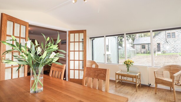 Ein Essbereich mit einem Holztisch, Stühlen und einer Blumenvase. Ein Fenster bietet Blick auf ein Steinhaus und einen Garten.