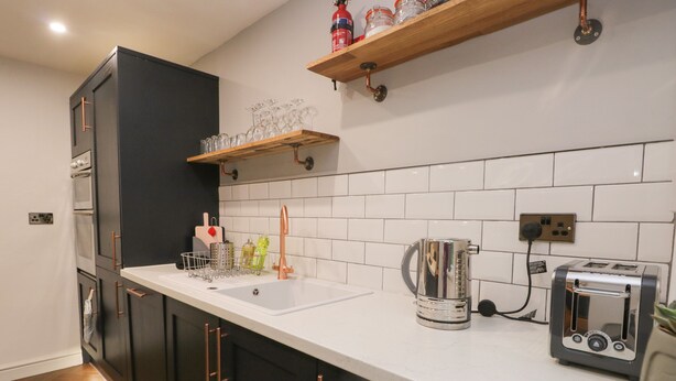 Une cuisine moderne avec un plan de travail blanc, des meubles noirs, une bouilloire en acier inoxydable, un grille-pain et une étagère en bois au-dessus de l’évier.
