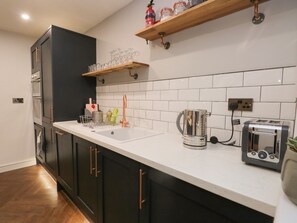 A modern kitchen with a white countertop, black cabinets, a stainless steel kettle, a toaster, and a wooden shelf above the sink. - Ravenglass Retreat (Ravenglass)