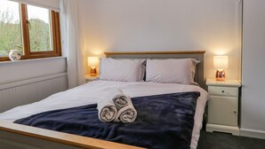 Un lit bien fait, avec une couverture bleue et des serviettes pliées, deux lampes de chevet, une fenêtre avec des rideaux et une petite table de chevet agrémentée d’un objet décoratif.