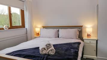 Cama bem arrumada com manta azul e toalhas dobradas, duas luminárias de cabeceira, janela com cortinas e uma mesinha de cabeceira com um objeto decorativo.