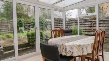 Uma varanda envidraçada com uma mesa de jantar e cadeiras, grandes portas de vidro e vista para um jardim.