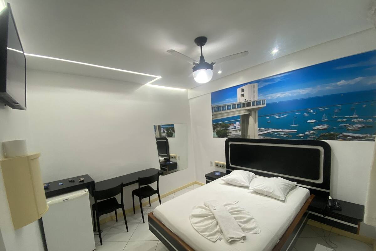 Un dormitorio con cama, escritorio, sillas, un refrigerador pequeño y un cuadro con vista al mar.