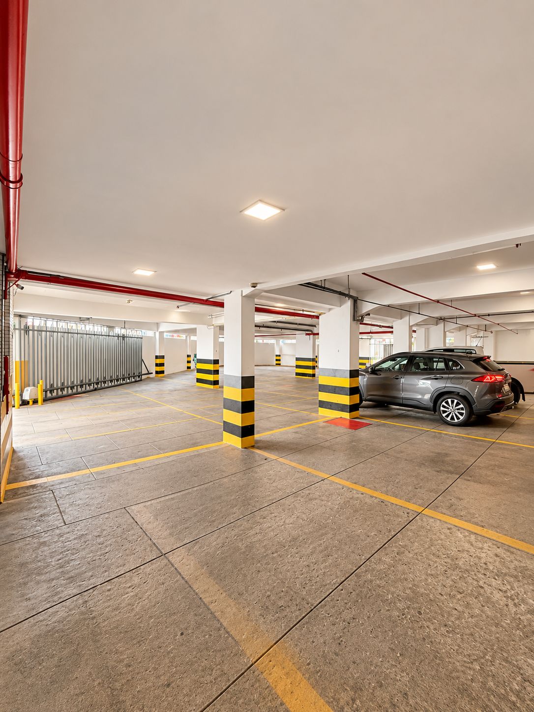 En parkeringsgarage med betongulv, søjler i gule og sorte striber og en grå bil parkeret.