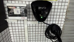 Dispositivo fijado a la pared con un cable conectado, una caja etiquetada 'STECK' con una etiqueta de certificación, y una pared con baldosas formando un patrón de cuadrícula.