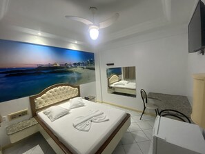 Ein Hotelzimmer mit Bett, Deckenventilator, großem Wandbild mit Strandmotiv, Spiegel und Wand-Fernseher.