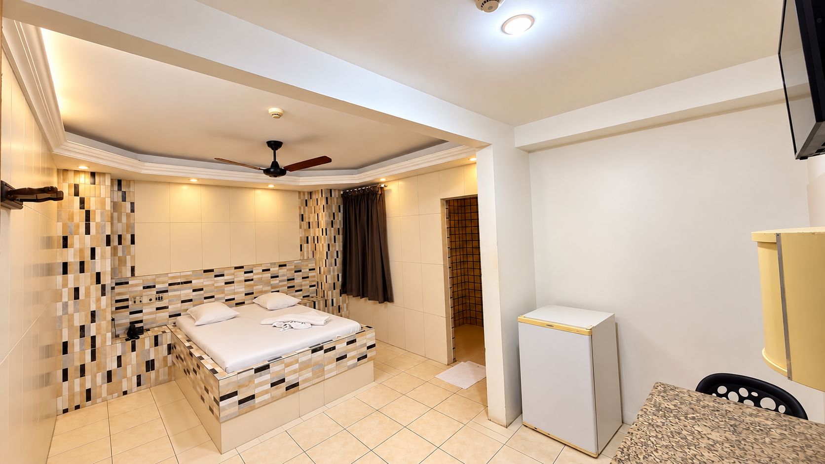 Habitación de hotel con cama, ventilador de techo, refrigerador pequeño y baño con ducha.