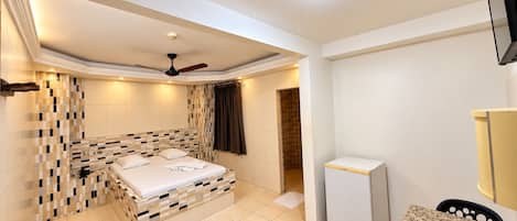 Habitación de hotel con cama, ventilador de techo, refrigerador pequeño y baño con ducha.