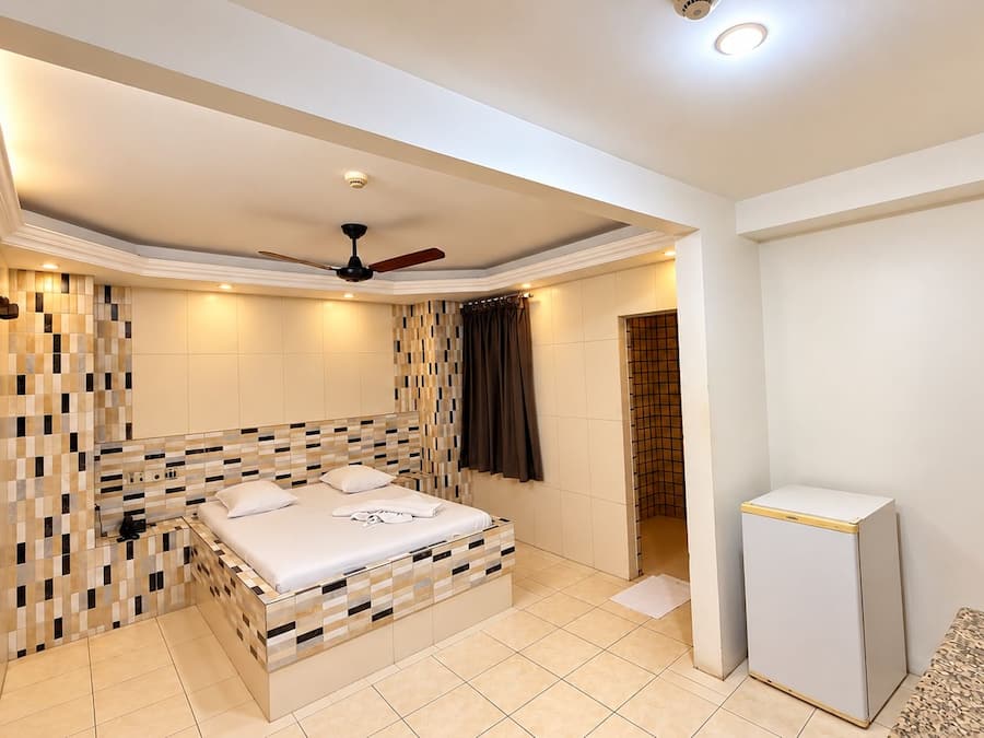 Ein Hotelzimmer mit einem Bett, einem Deckenventilator, einem kleinen Kühlschrank und einem Badezimmer mit Dusche.