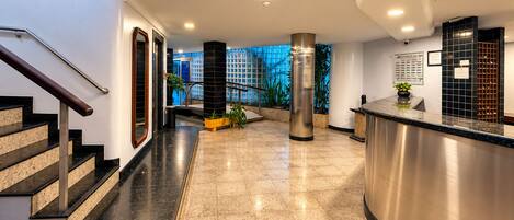 A hotel lobby with a reception desk, a staircase, and a view of the outside.
