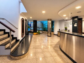 A hotel lobby with a reception desk, a staircase, and a view of the outside. - Hotel Novo Plano (Salvador)