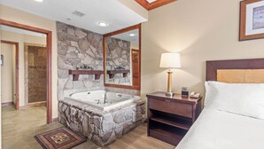 A hotel bathroom with a stone sink, a large mirror, a bathtub, and a bedside table with a lamp.