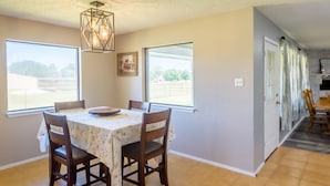 A dining room with a table set for four, large windows, and a kitchen visible in the background. - Holiday Decorations l Spacious l 1 Mile to Downtown Bandera l Dog Friendly (Bandera)