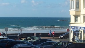 Uma rua costeira com carros estacionados, uma praia ao fundo e um edifício com uma placa que diz 'BAR Le Micro sillon'.