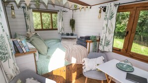 Ein gemütliches, helles Zimmer mit Bett, Schreibtisch und Stuhl. Auf dem Bett liegen Bücher, auf dem Schreibtisch steht eine Pflanze und es gibt ein Fenster mit Vorhängen.