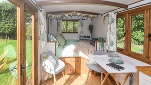 Ein gemütliches, helles Zimmer mit einem Bett, einem Tisch und einem Sessel. Ein Fenster mit Vorhängen, eine Pflanze und eine Pendelleuchte sind ebenfalls vorhanden.