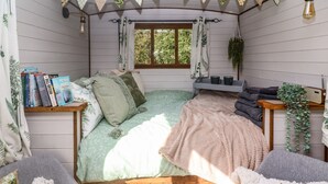 Ein gemütliches Schlafzimmer mit Bett, Nachttisch mit Büchern, Vorhängen am Fenster und einer hängenden Pflanze.