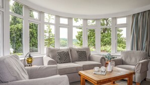 A cozy living room with a grey sofa, two armchairs, and a wooden coffee table. Large windows offer a view of greenery outside. - Ynys Castell (Menai Bridge)