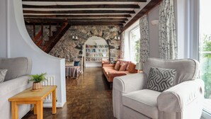 A cozy living room with a stone wall, wooden beams, a bookshelf, and a view through a window. - Ynys Castell (Menai Bridge)